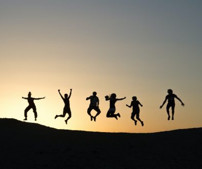 6 people jump in the air forming silhouettes on the top of a hill as the sun sets