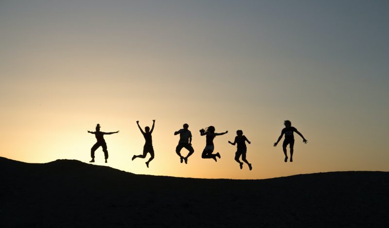 6 people jump in the air forming silhouettes on the top of a hill as the sun sets