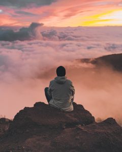 A man in a grey hoodie and a black beanie sits on a rock on top of a mountain overlooking a cloud inversion at sunrise or sunset