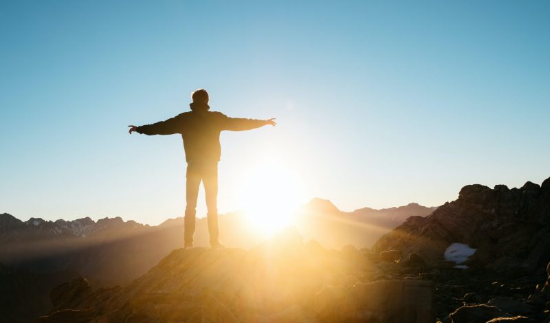 silhouette of a person in outdoor gear standing on a mountain as the golden light of the sun at dawn shines.